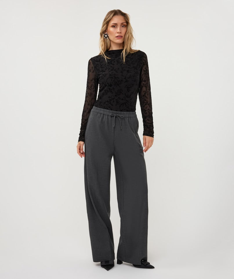 A woman modeling a black long-sleeve top with sheer lace sleeves and dark gray wide-leg drawstring pants against a plain light gray studio background.