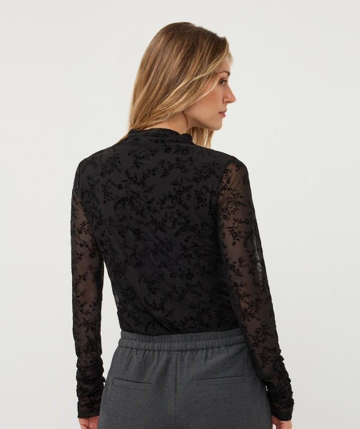 A woman shown from the back wearing a black sheer floral lace long-sleeve top and gray drawstring pants against a plain light gray studio background.