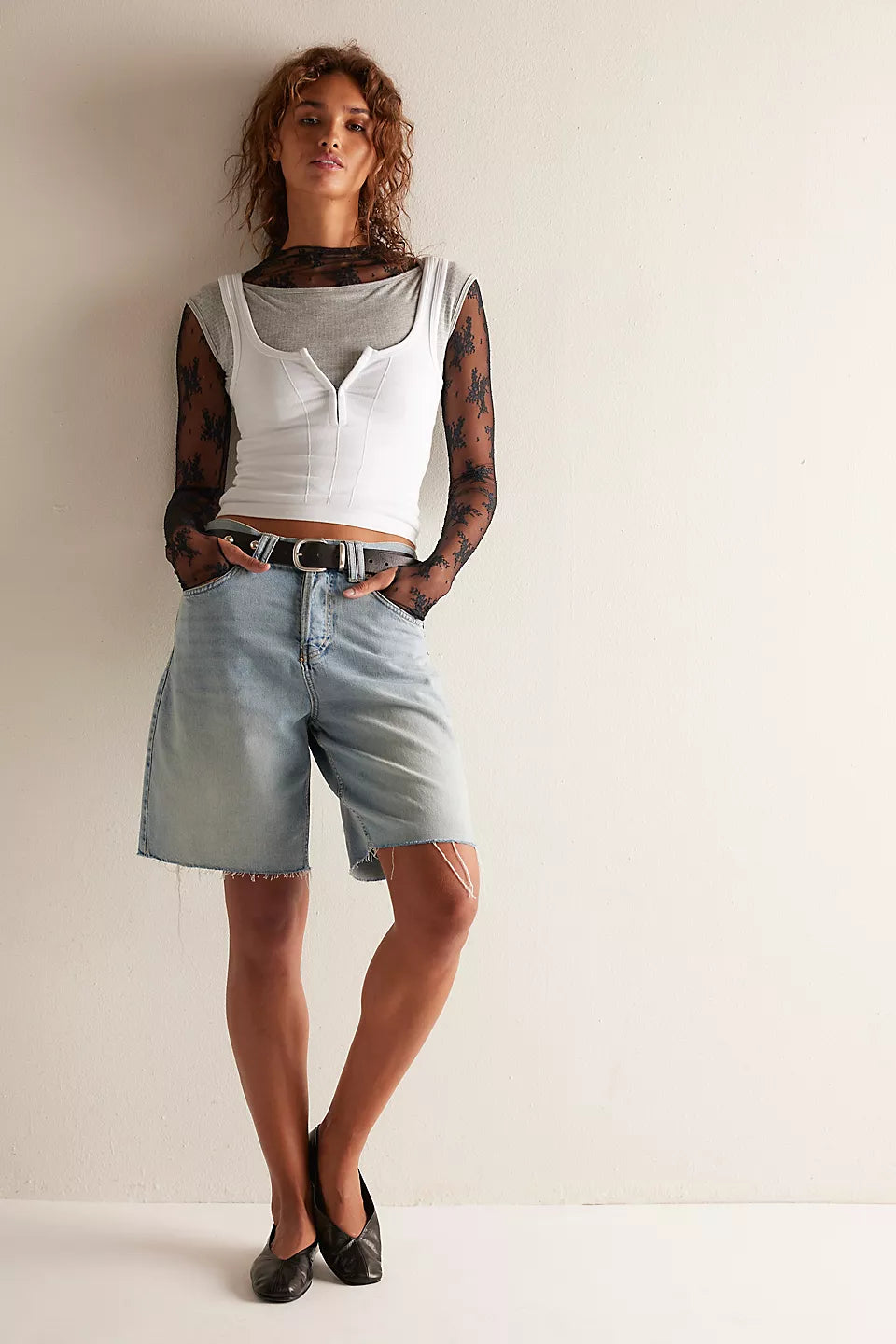 A model stands against a plain off-white wall wearing a white cropped tank top over a sheer black patterned long-sleeve shirt, light-wash denim shorts, and black flats.