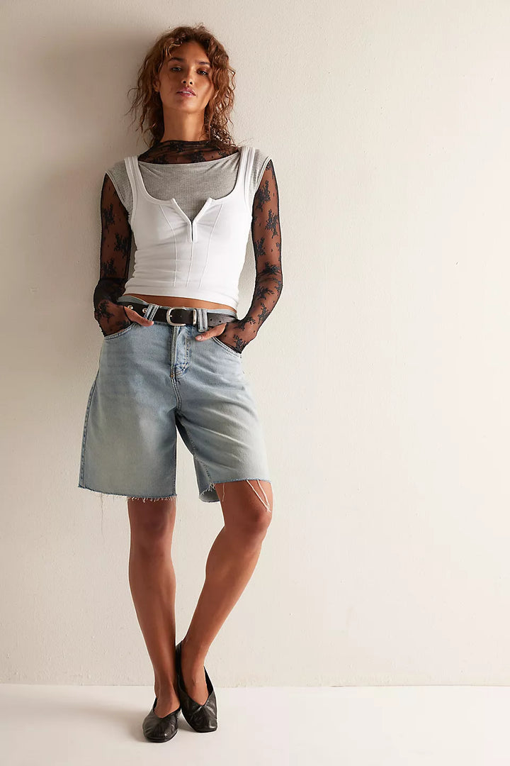 A model stands against a plain off-white wall wearing a white cropped tank top over a sheer black patterned long-sleeve shirt, light-wash denim shorts, and black flats.