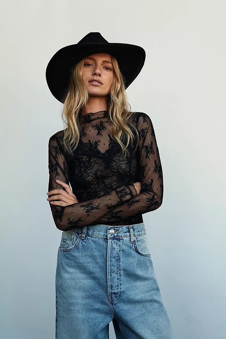 A woman wearing a black wide-brim hat, a sheer black floral lace long-sleeve top, and light blue high-waisted jeans, standing against a light gray studio background.