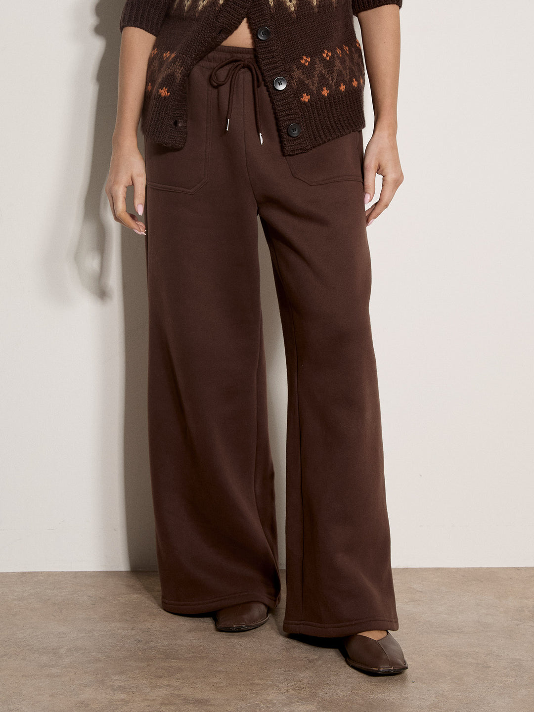 A person wearing dark brown wide-leg sweatpants with a drawstring waist, paired with a brown patterned cardigan and brown flats, standing against a plain off-white wall.