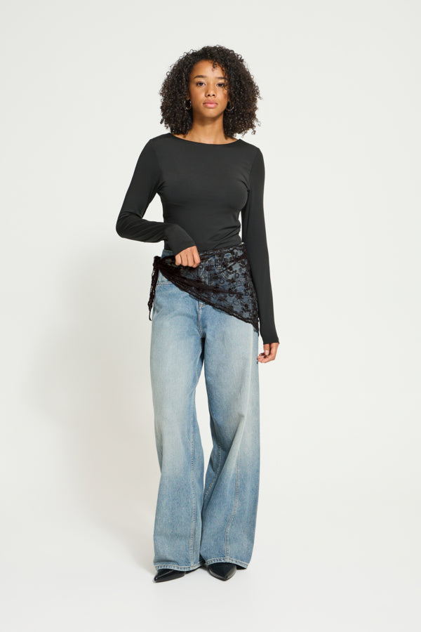 A model stands against a light gray studio background wearing a black long-sleeve top with a sheer black lace overlay, paired with wide-leg light-wash blue jeans and black shoes.