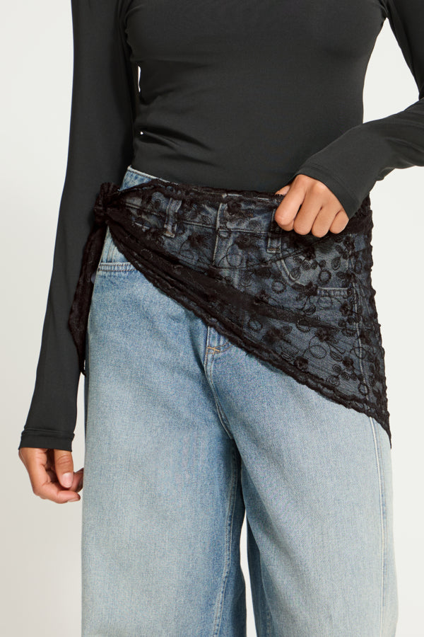 A person wearing a black lace wrap sash tied around the waist over a black long-sleeve top and light blue jeans, shown against a plain light background.