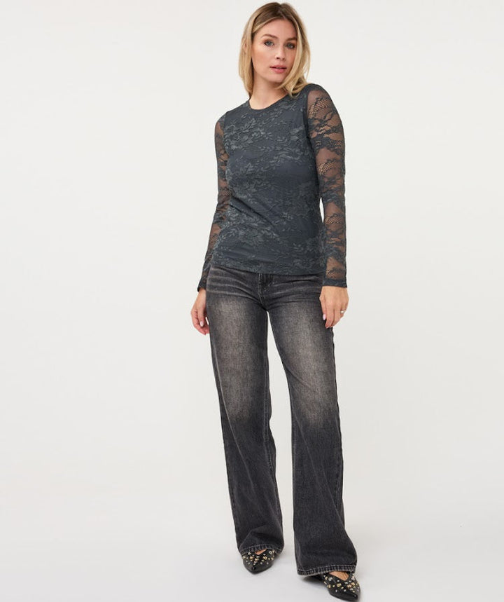 A woman modeling a dark charcoal gray long-sleeve top with sheer lace sleeves and dark gray wide-leg jeans, standing against a plain white studio background.