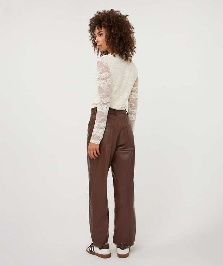 A model is shown from the back wearing brown wide-leg faux-leather trousers, a cream lace long-sleeve top, and white sneakers with brown soles, set against a plain white studio background.