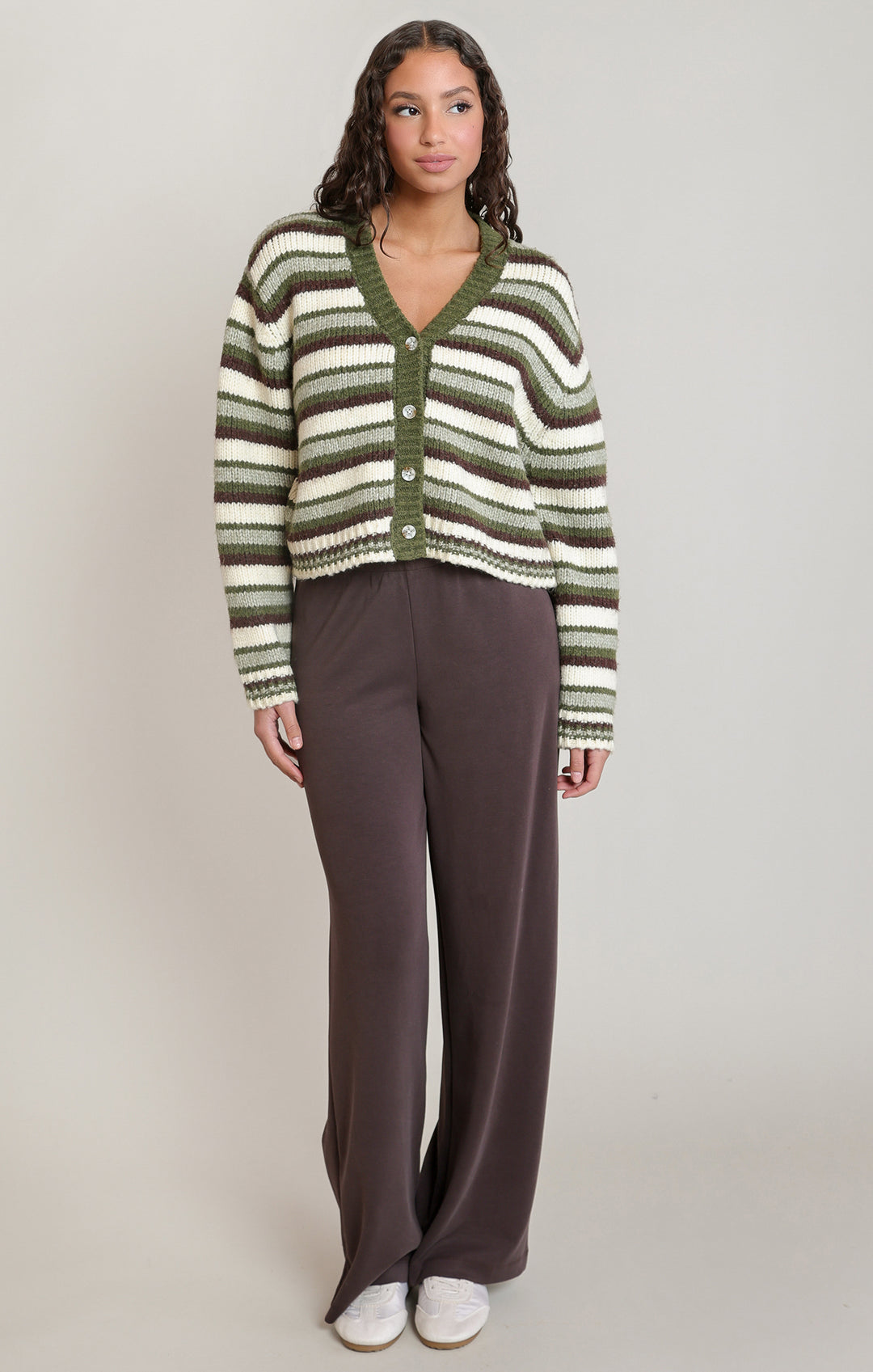 A woman modeling a cropped knit cardigan with horizontal stripes in olive green, cream, and brown, paired with dark brown wide-leg pants and white sneakers, against a plain light gray studio background.