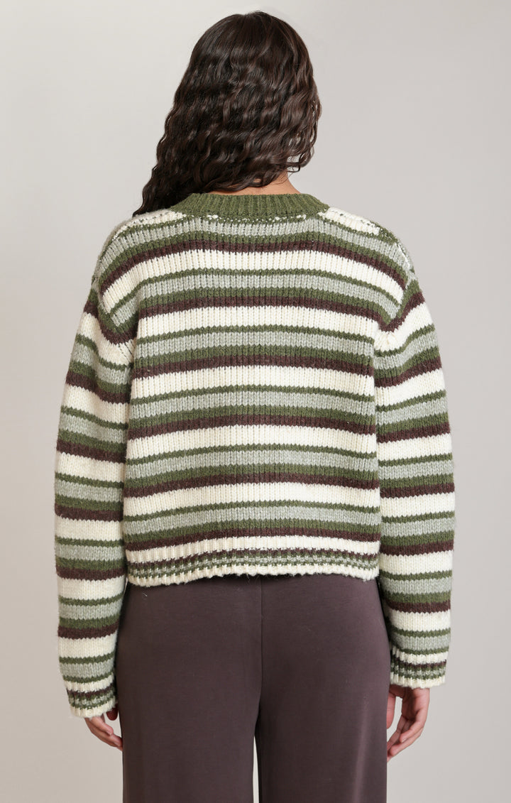 A woman viewed from the back wearing a cropped knit sweater with horizontal stripes in olive green, cream, and dark brown, paired with dark brown pants, against a solid light gray background.