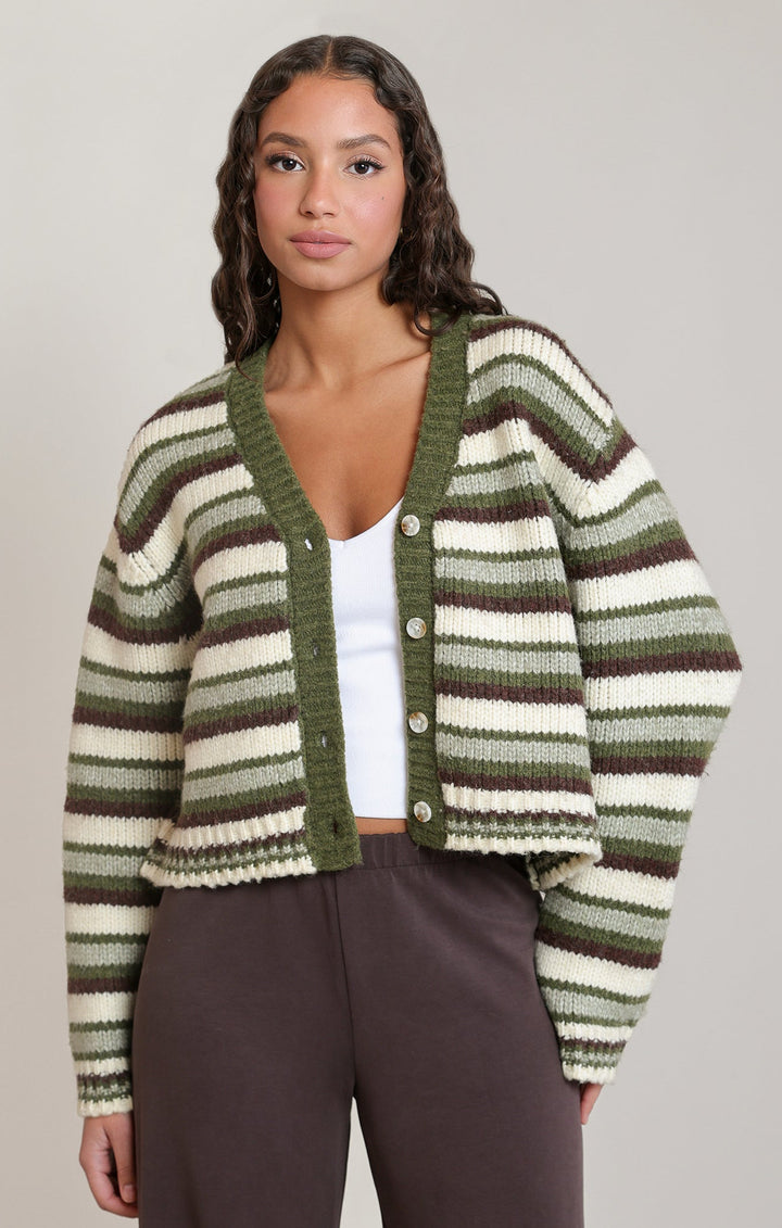 A woman modeling a cropped knit cardigan with horizontal stripes in olive green, dark brown, and cream, worn over a white top and dark brown pants against a plain light gray studio background.