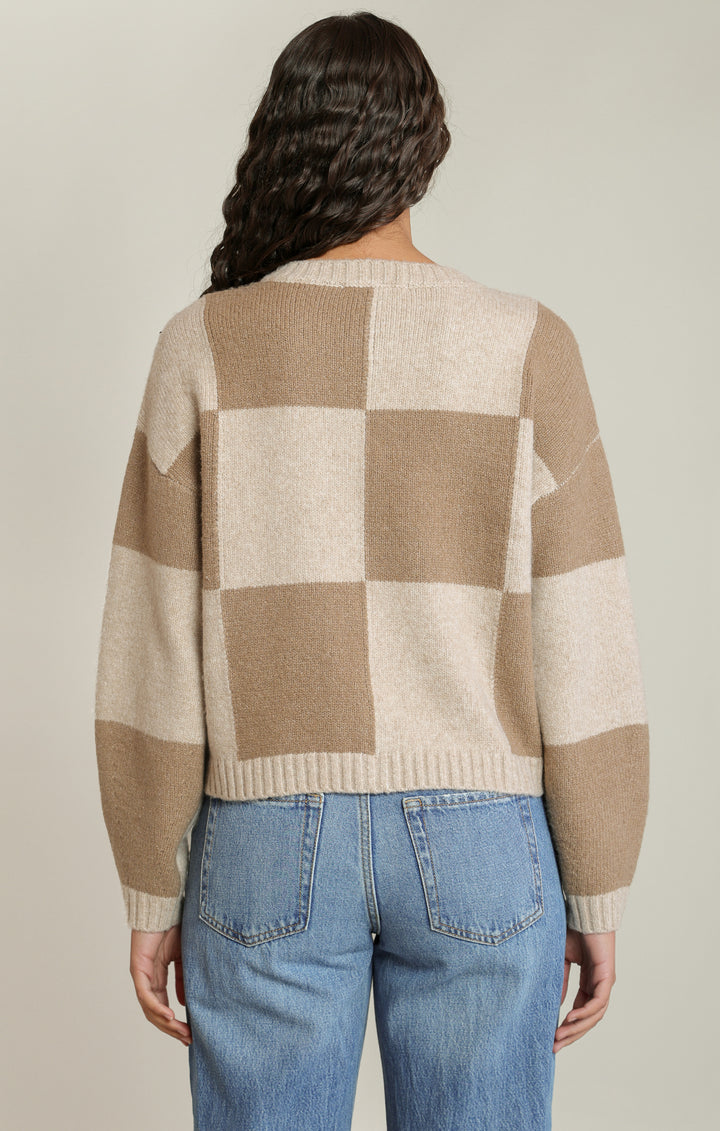 A woman viewed from the back wearing a beige and cream checkered knit sweater and blue jeans against a light gray studio background.