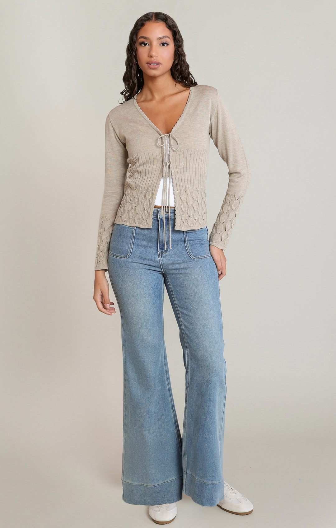 A woman modeling a beige cable-knit cropped cardigan with a tie-front detail, paired with light-wash blue flare jeans and white sneakers, against a plain light gray studio background.