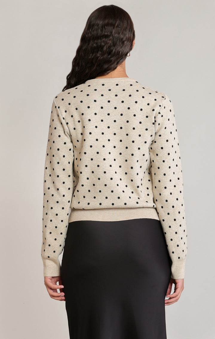 Woman wearing a beige sweater with black polka dots and black pants on a neutral background