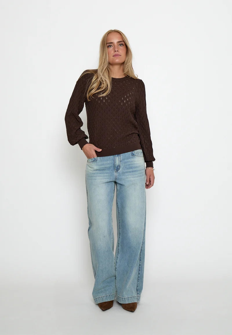 A woman modeling a dark brown long-sleeve knit sweater with a textured openwork pattern, paired with light-wash wide-leg jeans, standing against a plain white studio background.