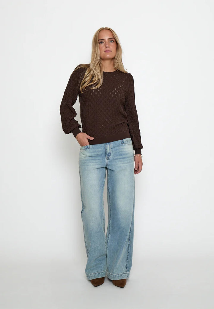 A woman modeling a dark brown long-sleeve knit sweater with a textured openwork pattern, paired with light-wash wide-leg jeans, standing against a plain white studio background.