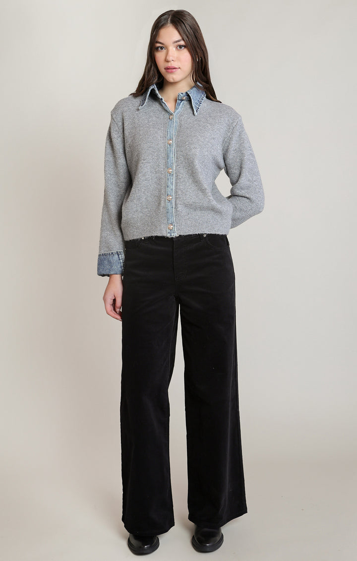 A woman stands against a plain light beige studio background wearing a light gray knit button-front cardigan over a light blue collared shirt, paired with black wide-leg pants and black shoes.