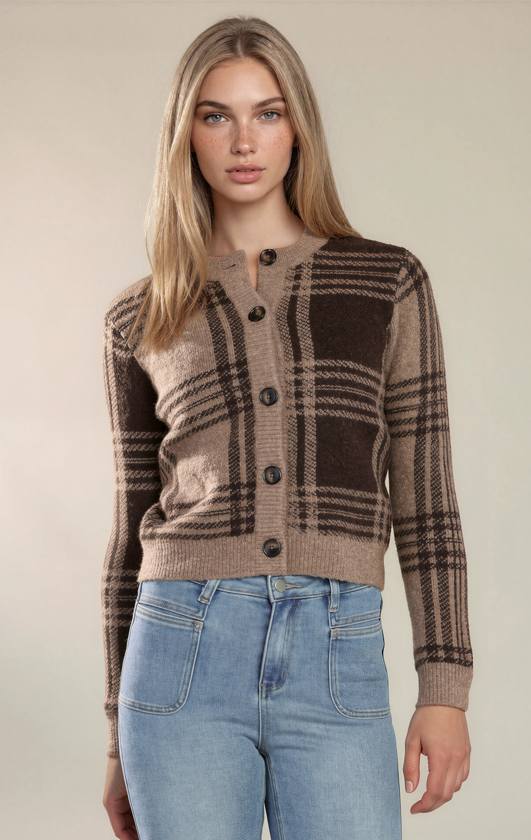 A woman modeling a brown and beige plaid button-up cardigan sweater with light blue jeans, set against a plain beige studio background.