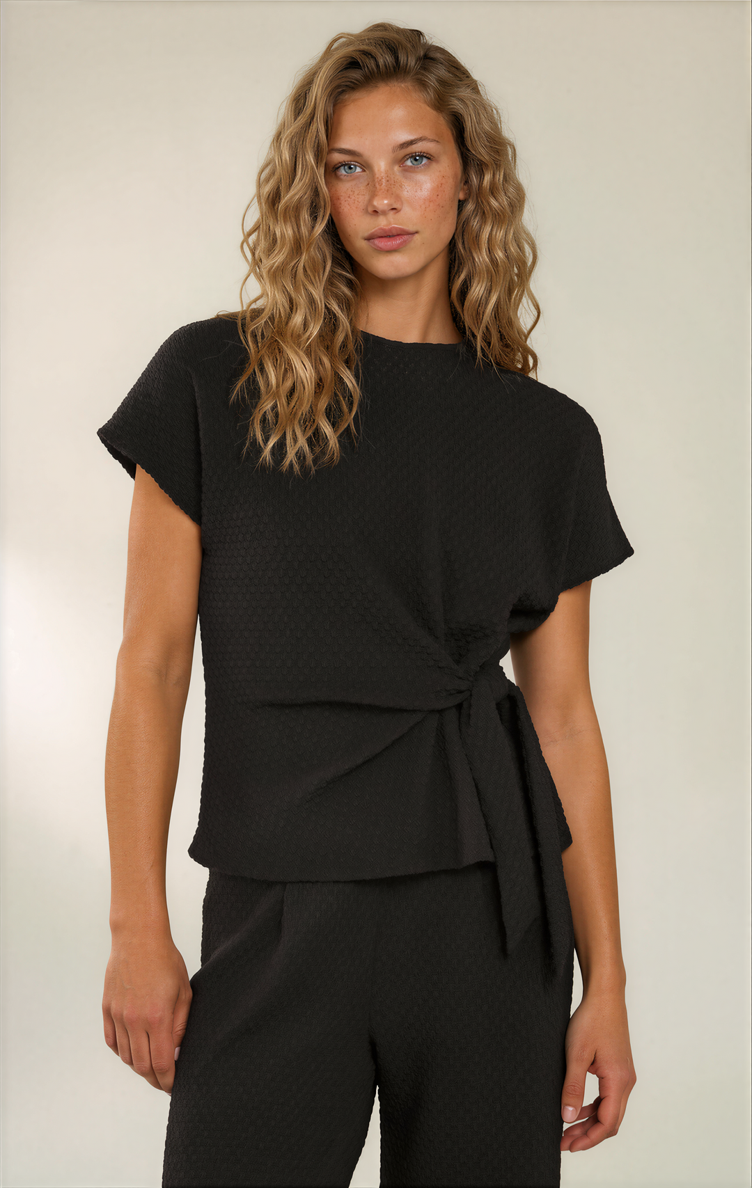 A woman modeling a black textured short-sleeve top with a twisted waist detail, paired with black trousers, against a plain light beige studio background.