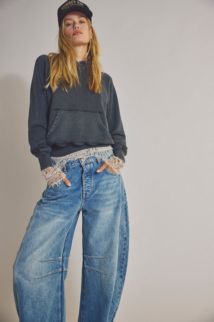 Person wearing a dark hoodie, lace gloves, and blue jeans against a plain background