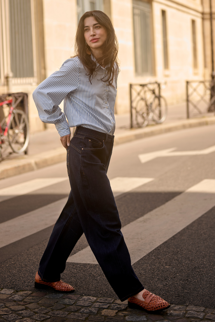 A woman walking across a city crosswalk wearing a light blue and white striped button-down shirt, dark wide-leg jeans, and brown woven loafers.