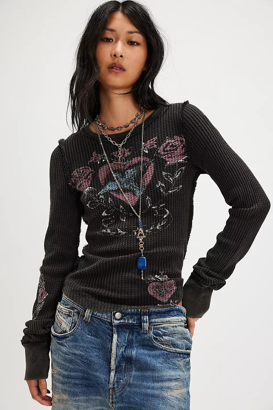 A model wearing a black ribbed long-sleeve top with a pink and white graphic design on the front, paired with blue jeans and layered necklaces, against a plain light gray background.