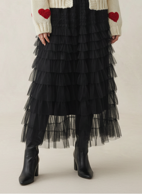 A woman stands against a light beige studio backdrop wearing a cream knit cardigan with red heart appliqués over a red top and a long black tiered tulle skirt, with black ankle boots.