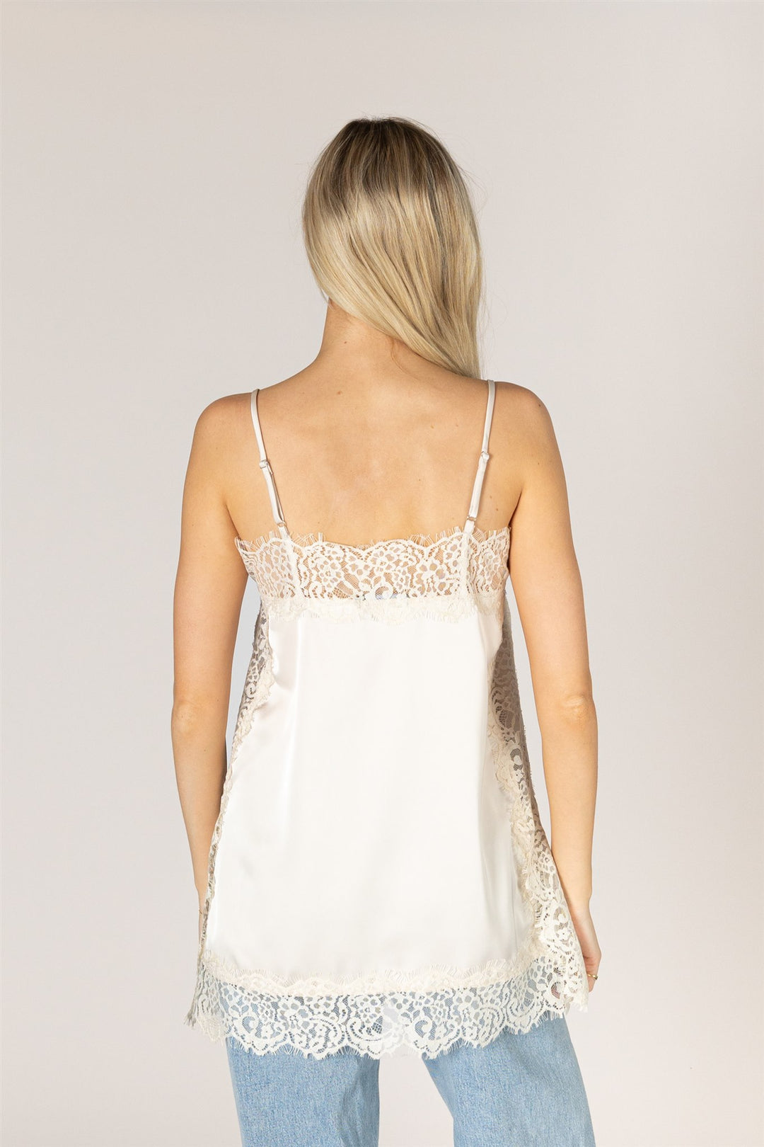 A woman viewed from the back wearing a white satin camisole top with delicate lace trim along the straps, neckline, and hem, paired with light blue jeans against a plain light gray studio background.
