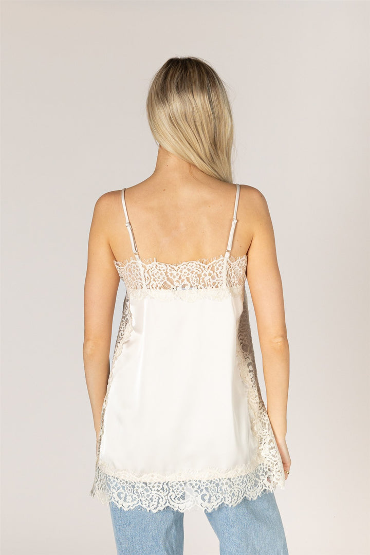 A woman viewed from the back wearing a white satin camisole top with delicate lace trim along the straps, neckline, and hem, paired with light blue jeans against a plain light gray studio background.