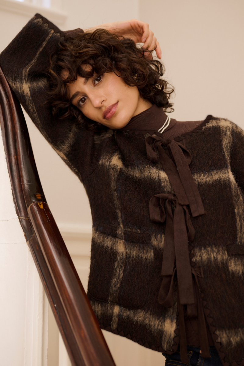 A woman with curly brown hair models a dark brown and beige plaid knit sweater with a high neck and front tie detail, leaning against a wooden banister in an indoor setting.