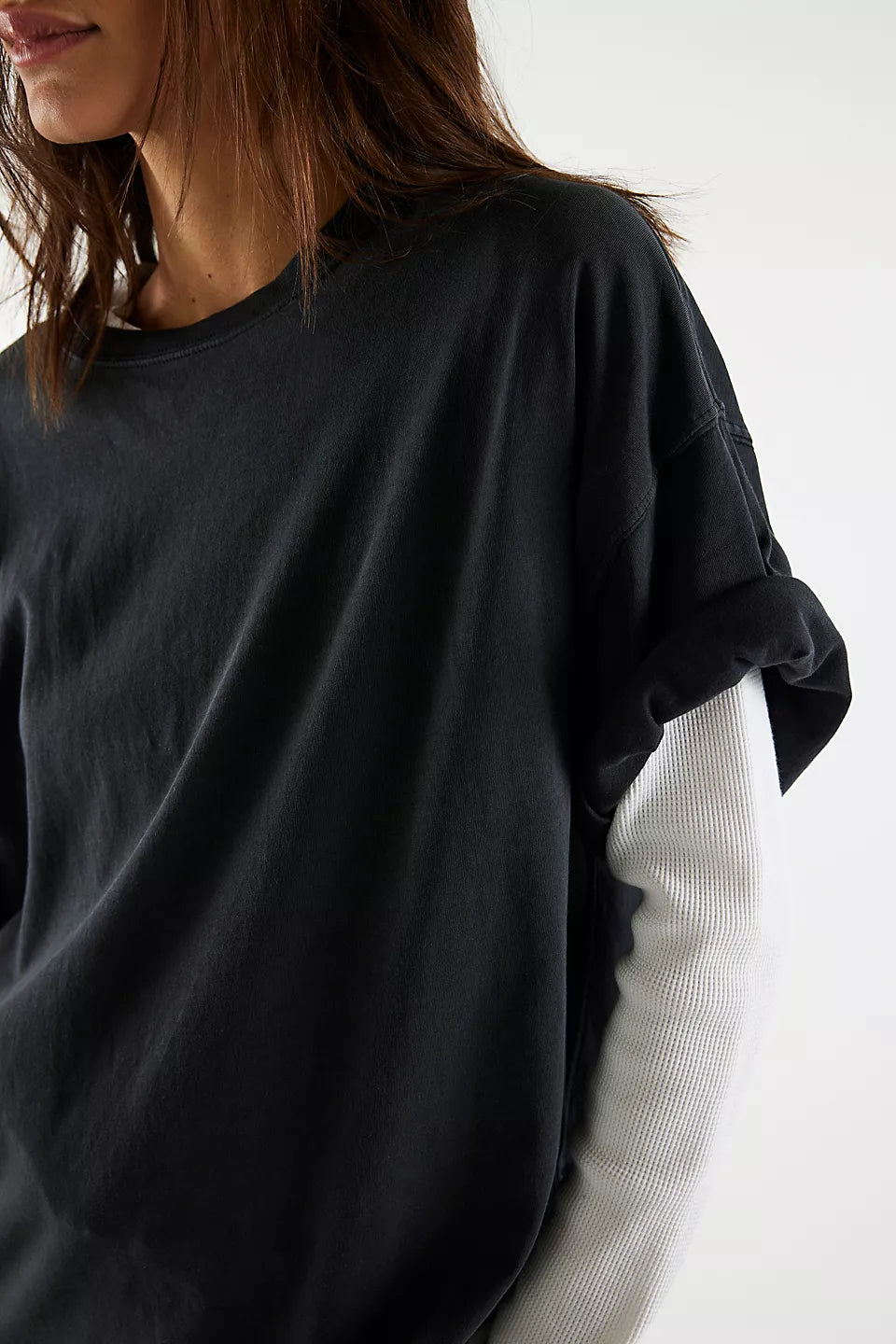 A close-up of a person wearing an oversized black long-sleeve top over a white ribbed long-sleeve shirt, against a plain light background.