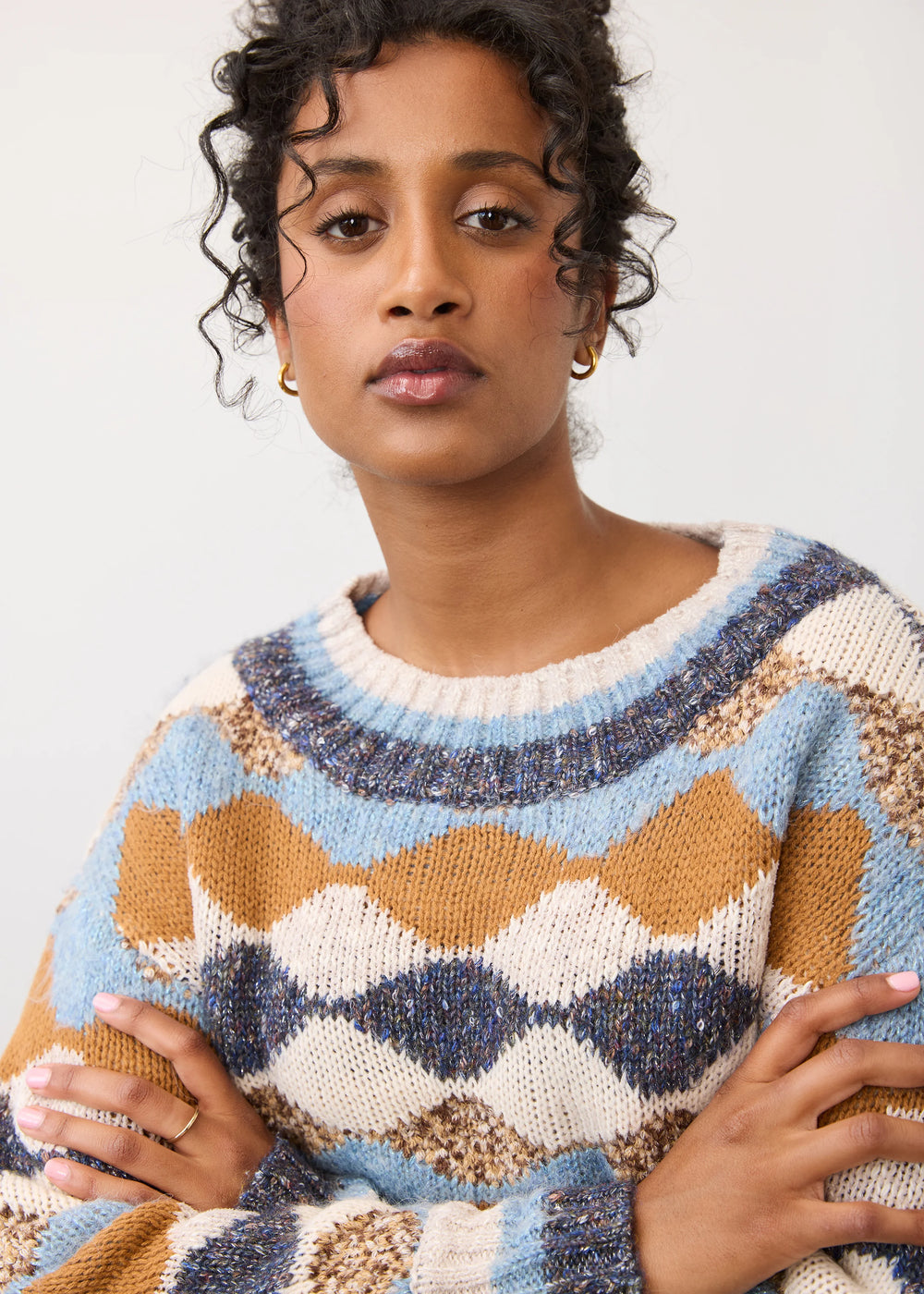 A woman wearing a chunky knit crewneck sweater with a geometric argyle pattern in light blue, mustard brown, cream, and navy blue, posed against a plain off-white background.