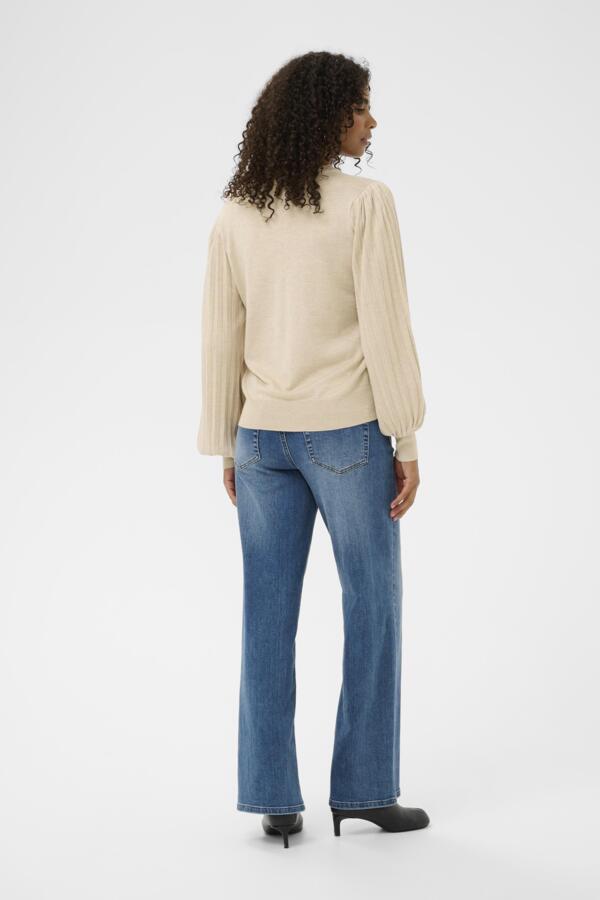 A woman with curly dark hair is shown from the back wearing a cream-colored ribbed knit sweater with balloon sleeves and blue jeans, set against a plain light gray studio background.