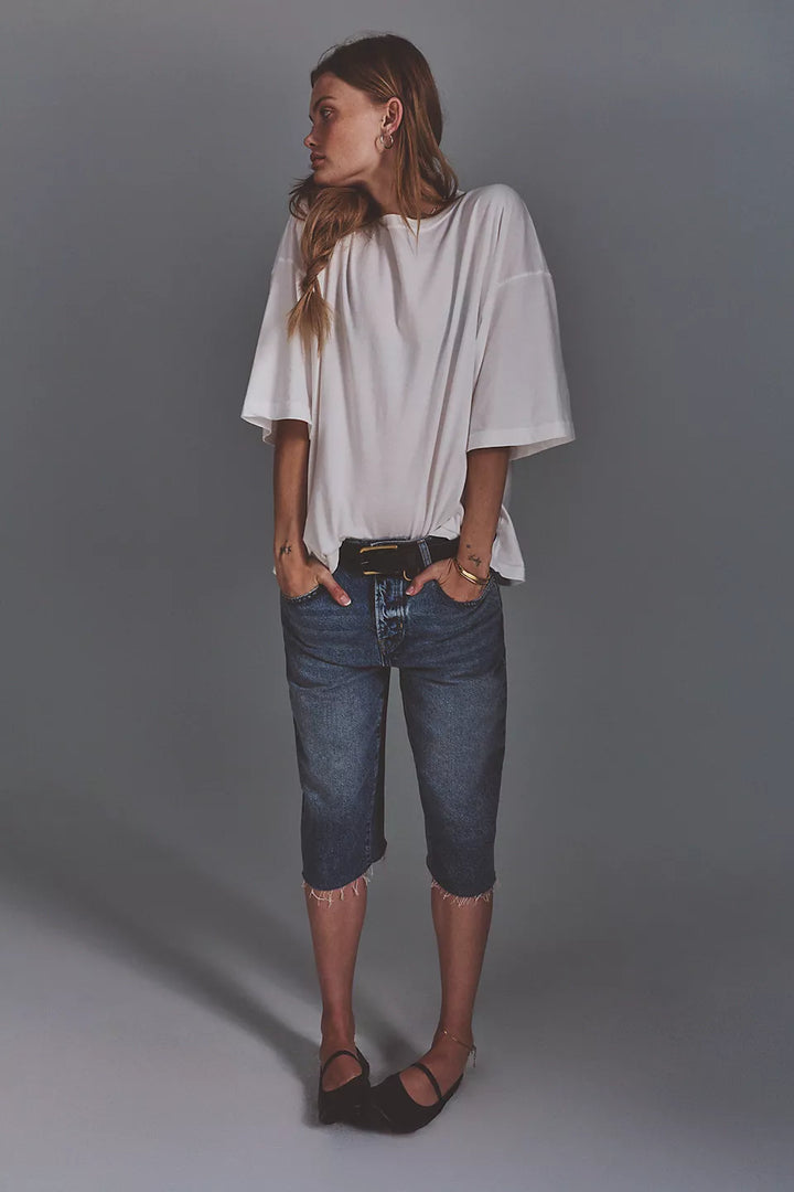A woman modeling a white short-sleeve top with wide bell sleeves, paired with dark blue cropped denim jeans and brown flat shoes, standing against a solid gray studio background.