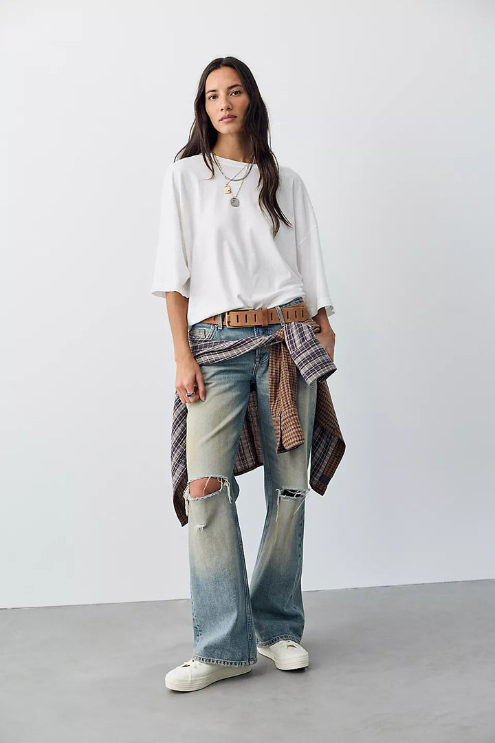 A woman modeling a white short-sleeve t-shirt, light-wash distressed flare jeans with knee rips, a brown belt, and a brown plaid shirt tied around her waist, paired with white sneakers against a plain studio background.