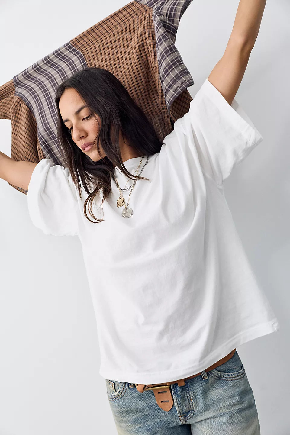 A woman wearing an oversized white short-sleeve t-shirt and light-wash blue jeans, holding a brown and beige plaid shirt over her shoulder against a plain light gray background.