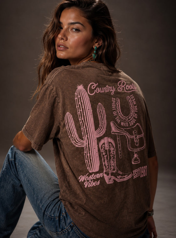 Woman wearing a brown t-shirt with a cactus and boot design on the back, sitting against a gray background.