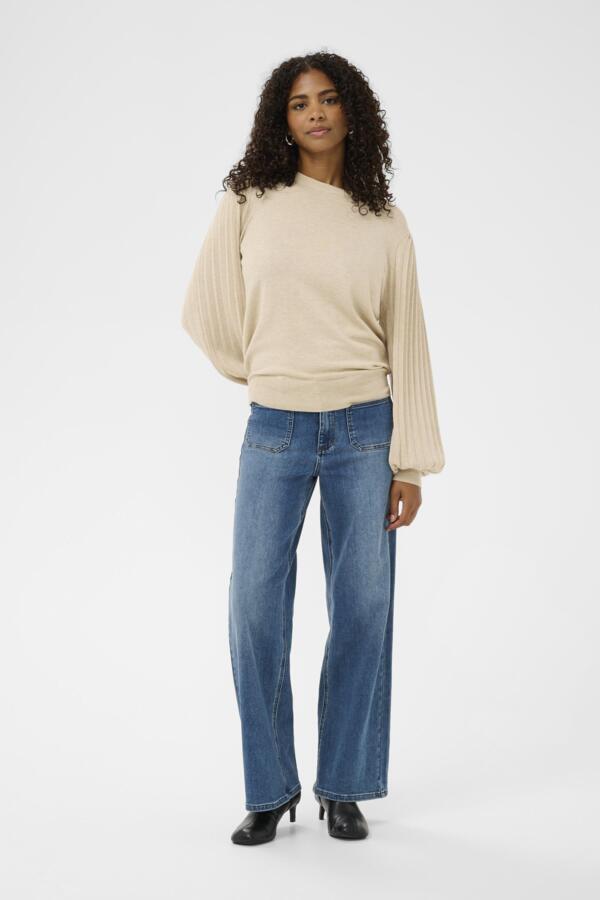 A woman with curly dark hair wearing a beige long-sleeve knit sweater with balloon sleeves and blue jeans, standing against a plain light gray studio background.
