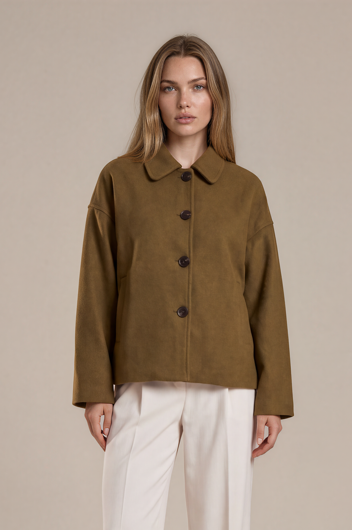 A woman stands against a plain beige studio background wearing a cropped olive-brown suede jacket with dark buttons, off-white wide-leg trousers, and black pointed heels.