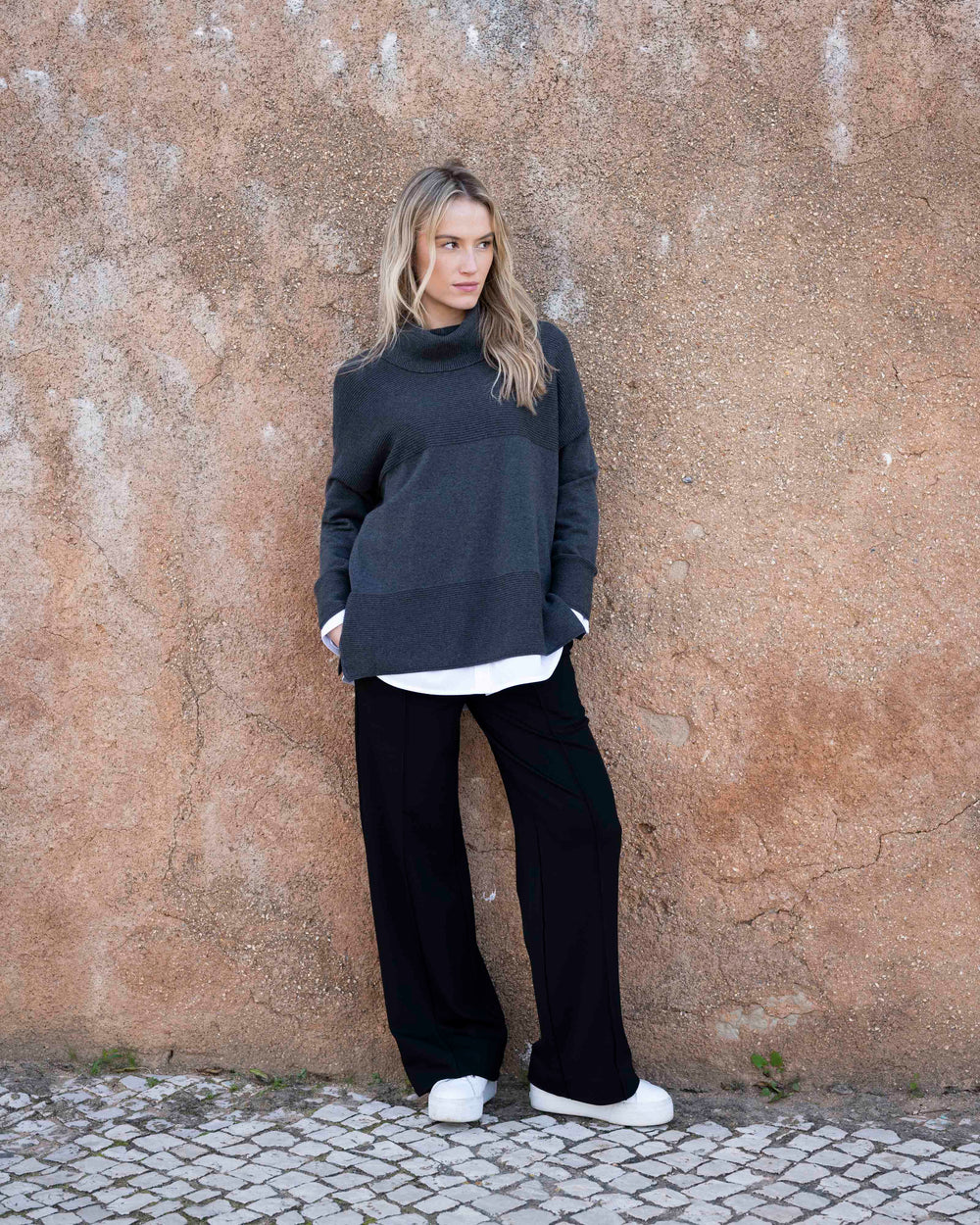 Woman wearing a dark gray sweater and black pants against a textured wall.