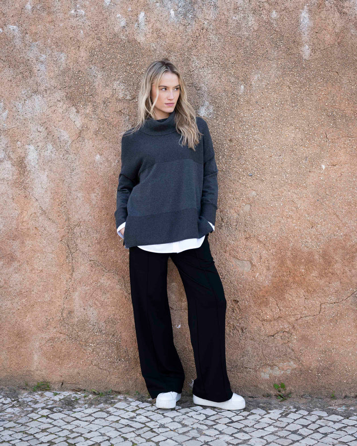 Woman wearing a dark gray sweater and black pants against a textured wall.