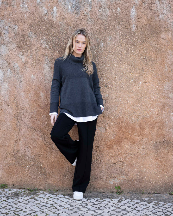 Woman wearing a dark sweater, white shirt, black pants, and white shoes against a textured wall.