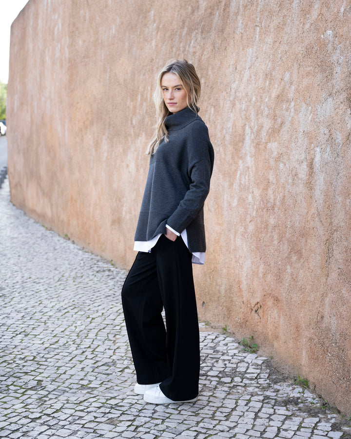 Woman wearing a dark sweater and black pants standing against a textured wall.