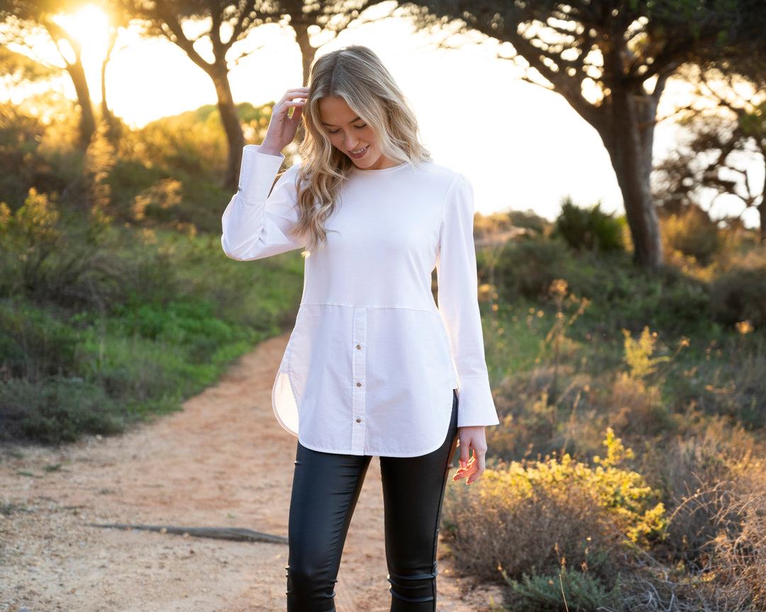 Woman in a white shirt and black pants standing on a dirt path with trees and grass in the background