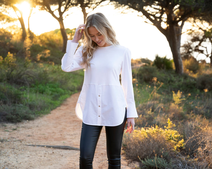 Woman in a white shirt and black pants standing on a dirt path with trees and grass in the background