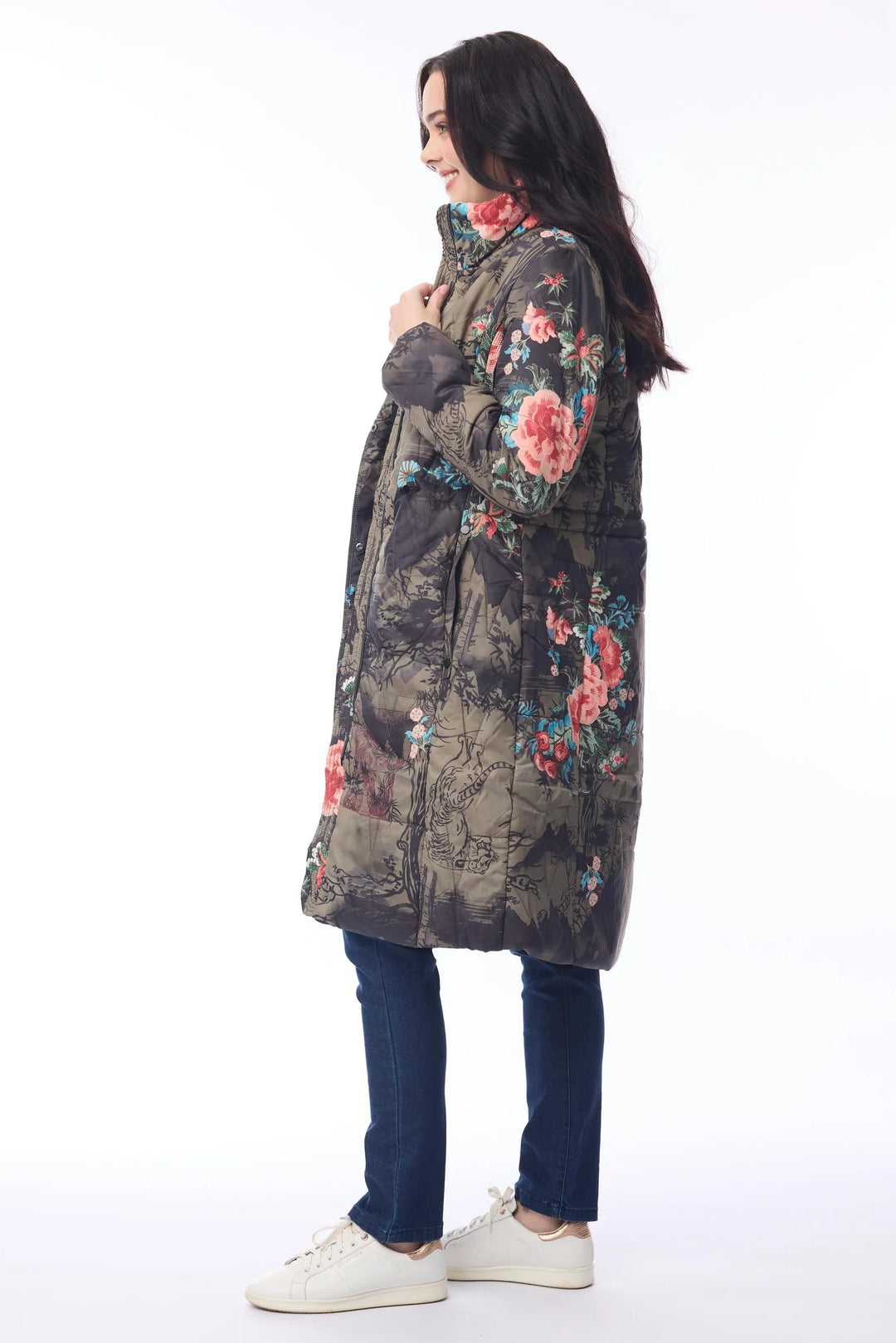 A woman wearing a long quilted puffer coat with a dark floral pattern, blue jeans, and white sneakers, standing in profile against a plain white studio background.