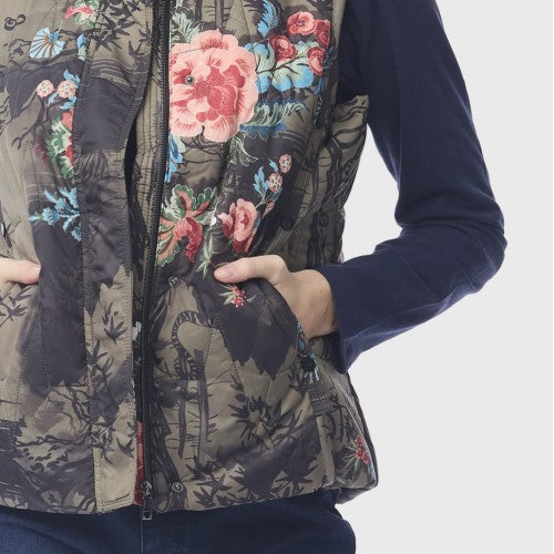 A close-up of a person wearing a quilted vest with a floral and bird print over a dark navy long-sleeve top. The vest features pink, red, and blue flowers with green leaves on a dark background, and the person&