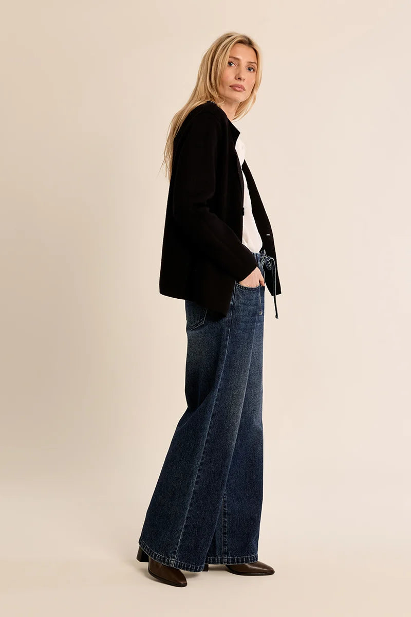 A woman modeling a black cropped blazer, wide-leg blue jeans, and dark brown shoes against a solid beige studio background.