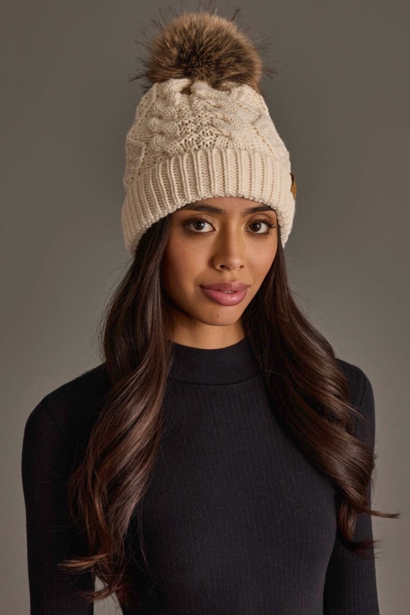 Woman wearing a beige knitted beanie with a pom-pom against a gray background