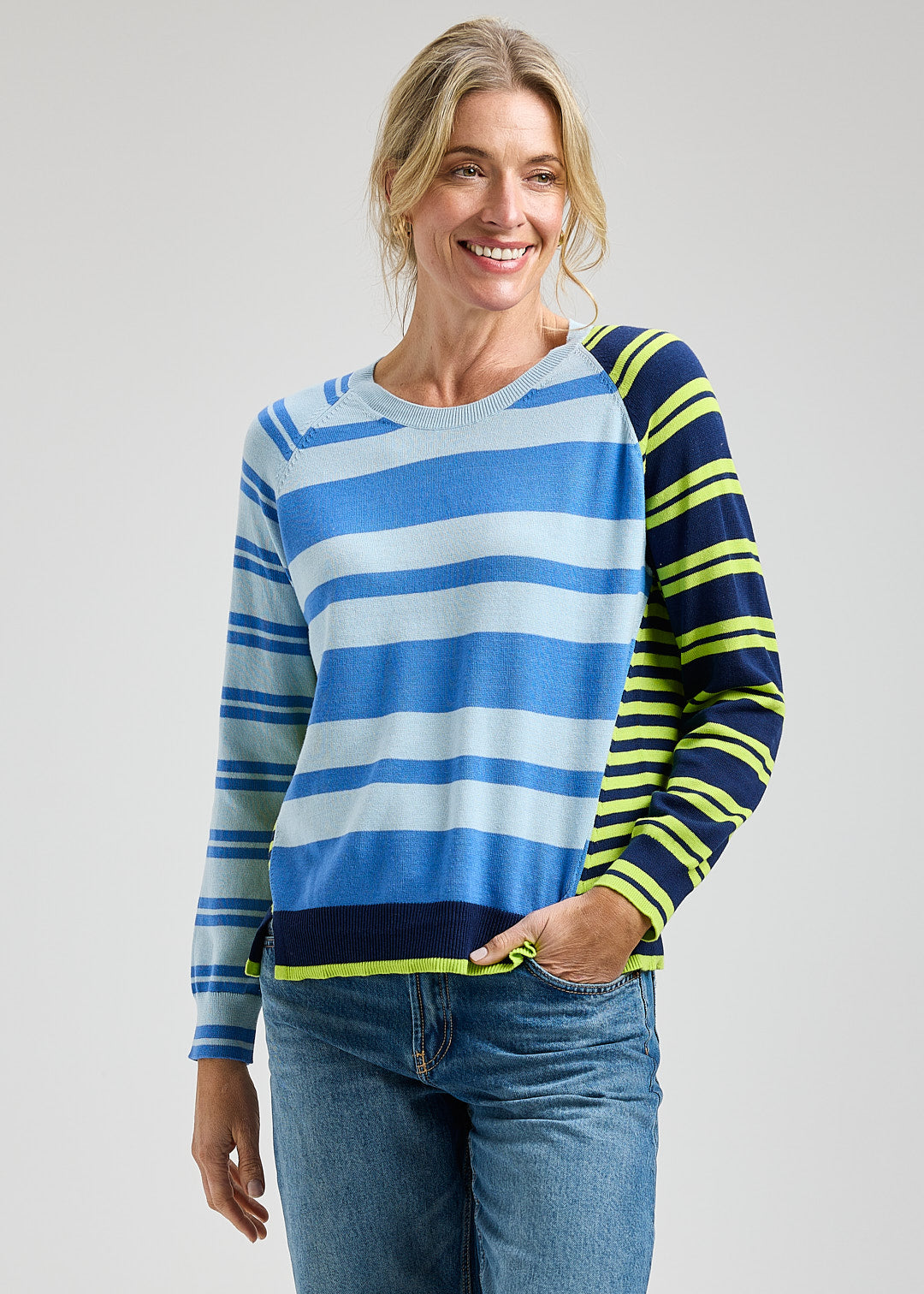 Woman wearing a blue, white, and green striped sweater with jeans on a plain background
