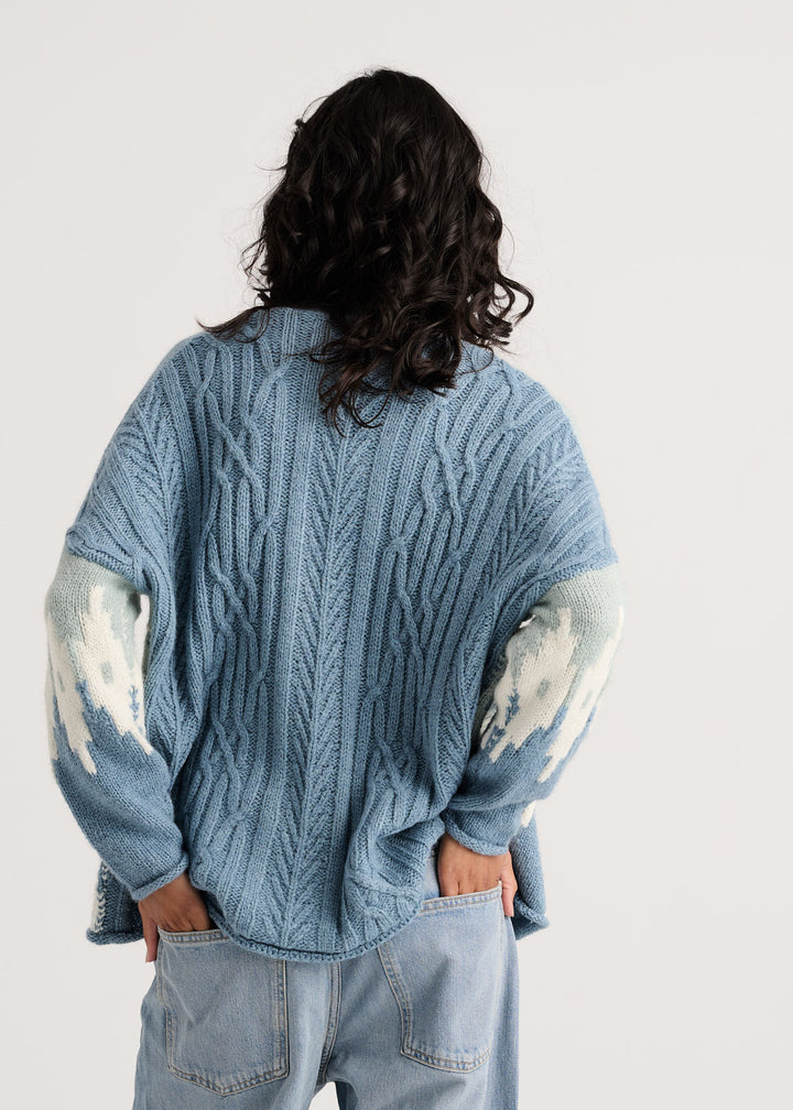 A person shown from the back wearing an oversized light blue cable-knit sweater with distressed white patches on the sleeves, paired with light-wash jeans, against a plain light background.