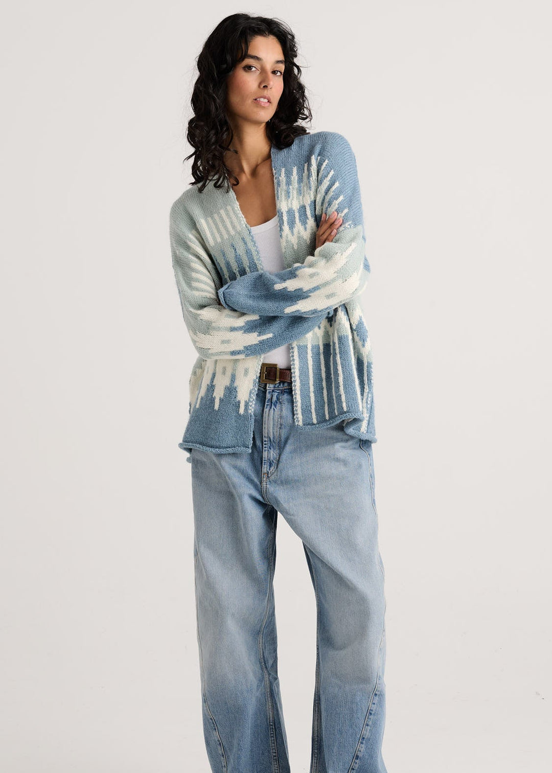 A woman stands against a light gray studio background wearing a light blue and white patterned cardigan, light-wash wide-leg jeans, and dark brown heeled boots.